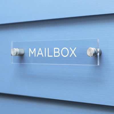 A custom Mailbox Acrylic sign from Girl Friday Shop, featuring crisp white letters, adorns a light blue wall elegantly mounted with two metal posts.