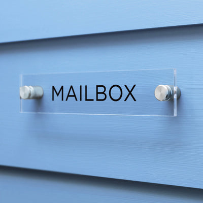 A custom-designed Mailbox Acrylic sign by Girl Friday Shop features MAILBOX in black letters on a sleek surface, mounted on a light blue wall with two chrome bolts.
