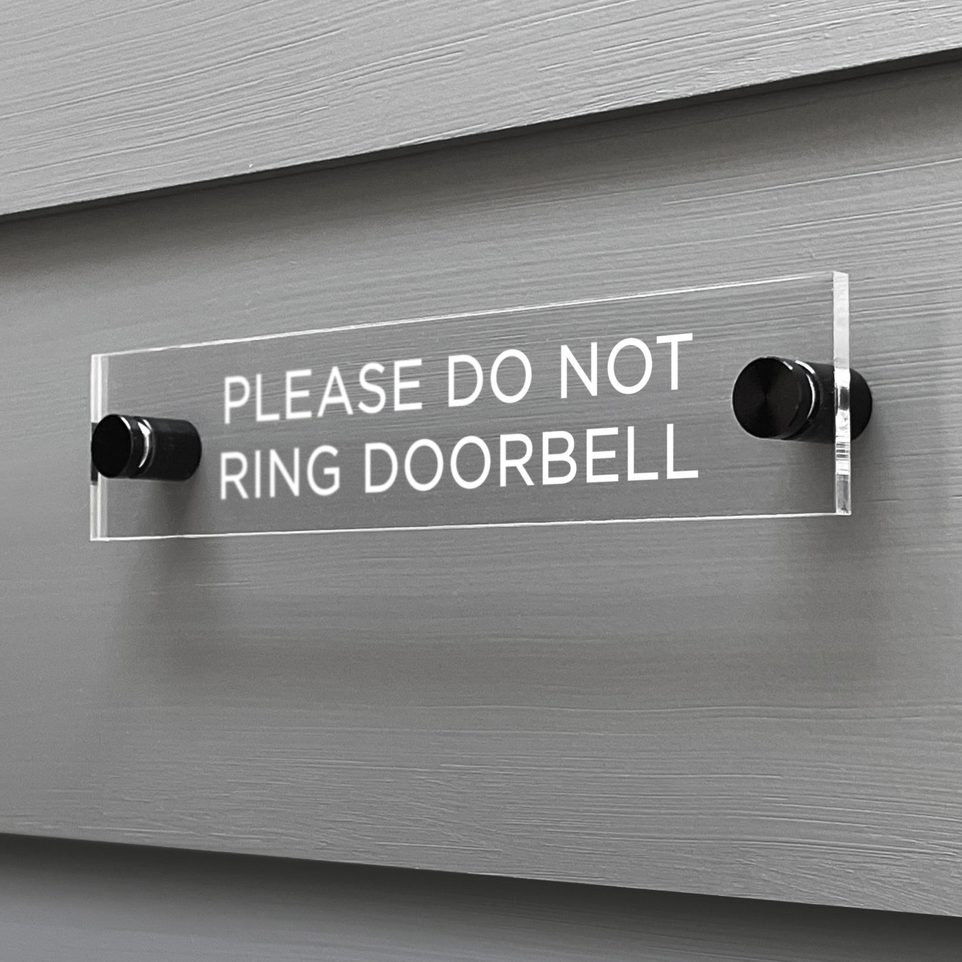 The Girl Friday Shops Please Do Not Ring Door Bell Acrylic Sign, secured with two black mounts, is displayed on a gray wall and features white lettering.