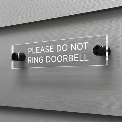 The Girl Friday Shops Please Do Not Ring Door Bell Acrylic Sign, secured with two black mounts, is displayed on a gray wall and features white lettering.
