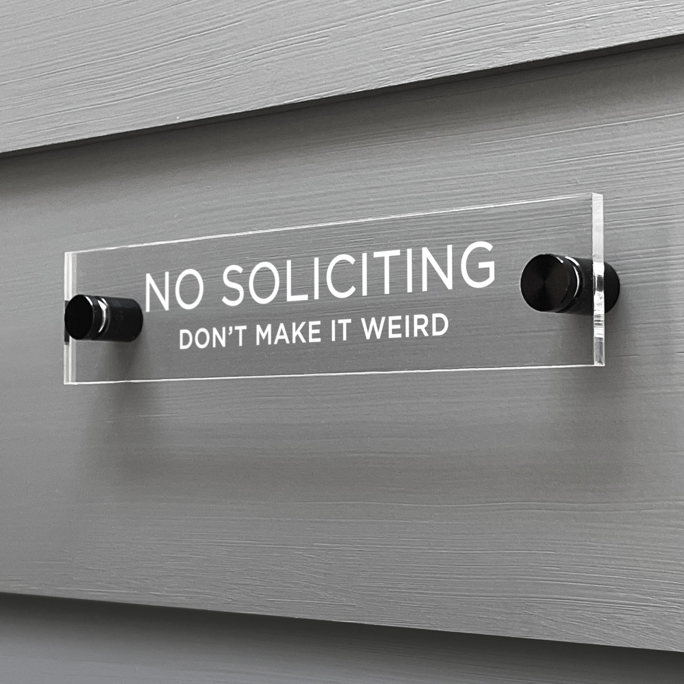 The Girl Friday Shops No Soliciting Dont Make It Weird Acrylic Sign is mounted on a gray wall with bold white letters and stylishly held by two round black posts.