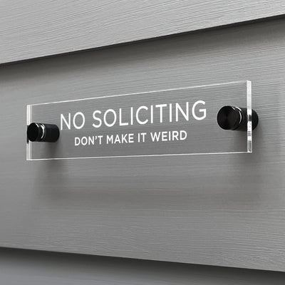 The Girl Friday Shops No Soliciting Dont Make It Weird Acrylic Sign is mounted on a gray wall with bold white letters and stylishly held by two round black posts.