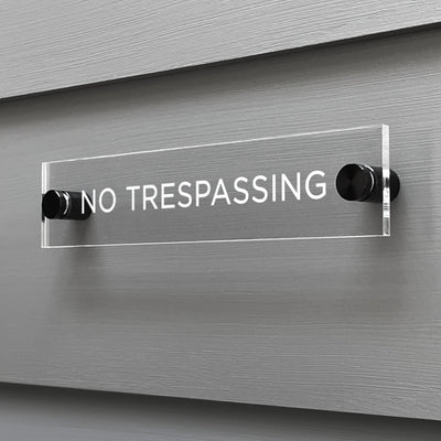 The sleek Girl Friday Shop No Trespassing Acrylic Sign, featuring a custom design, is mounted on a gray wall with stylish black metal fixtures.