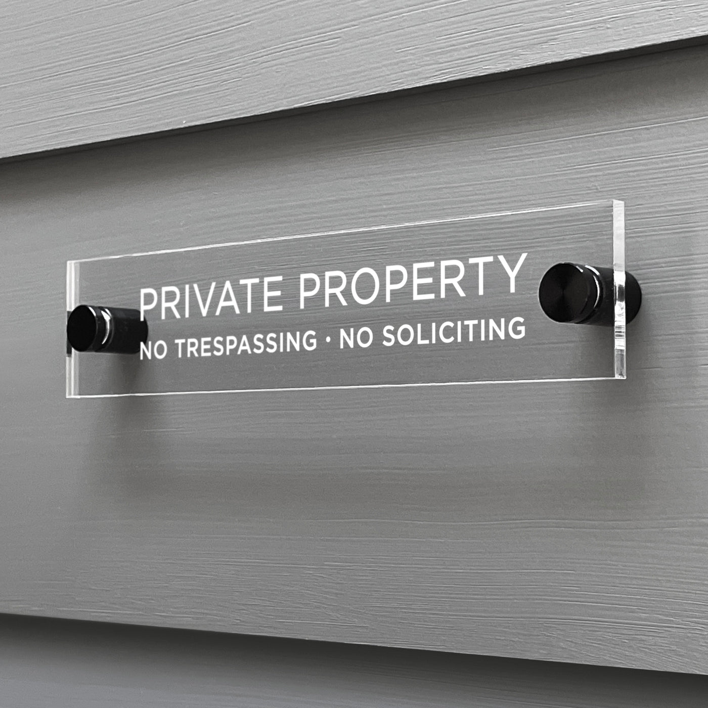 The Private Property Acrylic Sign by Girl Friday Shop features bold white text: PRIVATE PROPERTY - NO TRESPASSING - NO SOLICITING. Mounted on a gray wall with black standoffs, its sleek design ensures the message is clear and unmistakable.