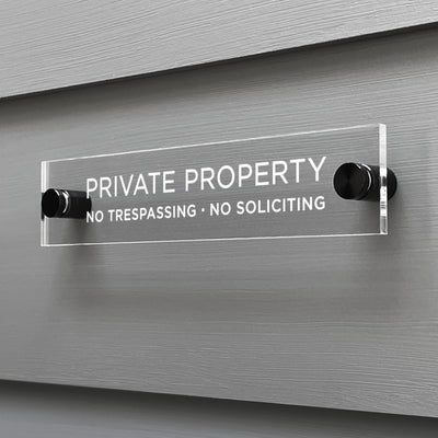The Private Property Acrylic Sign by Girl Friday Shop features bold white text: PRIVATE PROPERTY - NO TRESPASSING - NO SOLICITING. Mounted on a gray wall with black standoffs, its sleek design ensures the message is clear and unmistakable.