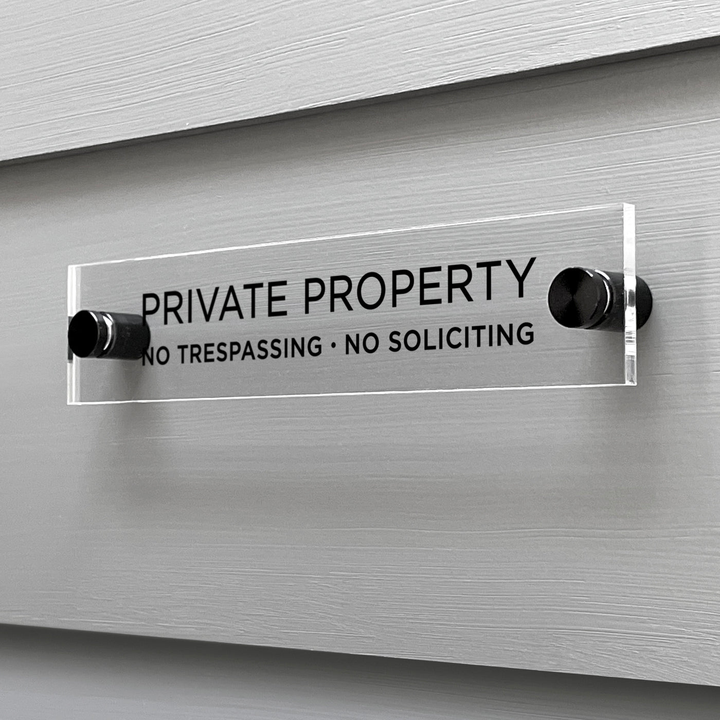 The Private Property Acrylic Sign by Girl Friday Shop is mounted on a gray wall, boldly displaying Private Property with the words No Trespassing · No Soliciting beneath. Its securely held by two black mounts.