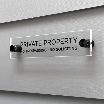 The Private Property Acrylic Sign by Girl Friday Shop is mounted on a gray wall, boldly displaying Private Property with the words No Trespassing · No Soliciting beneath. Its securely held by two black mounts.