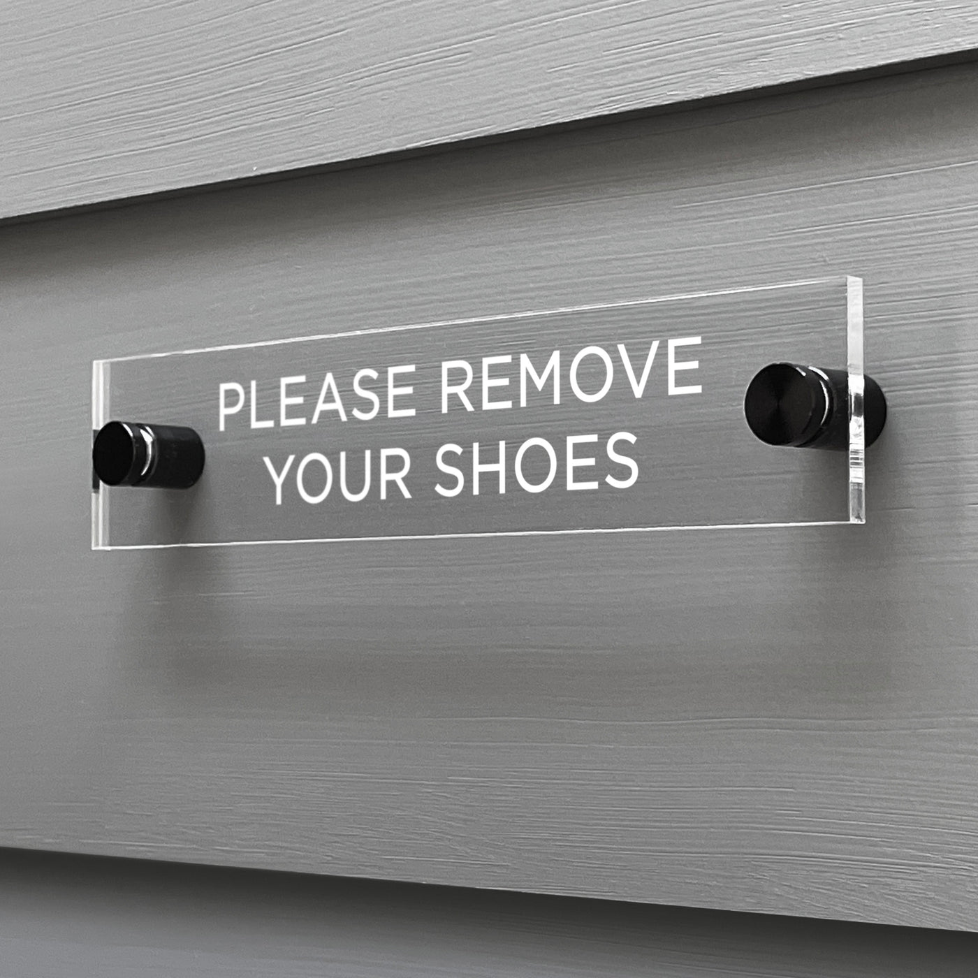 The Girl Friday Shops Please Remove Your Shoes Acrylic Sign features a clear design with black mounting posts, showcasing the text in white against a gray wall backdrop.