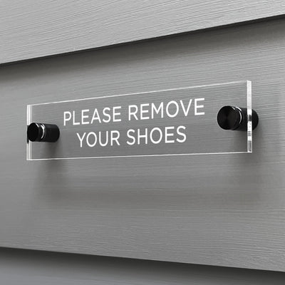 The Girl Friday Shops Please Remove Your Shoes Acrylic Sign features a clear design with black mounting posts, showcasing the text in white against a gray wall backdrop.