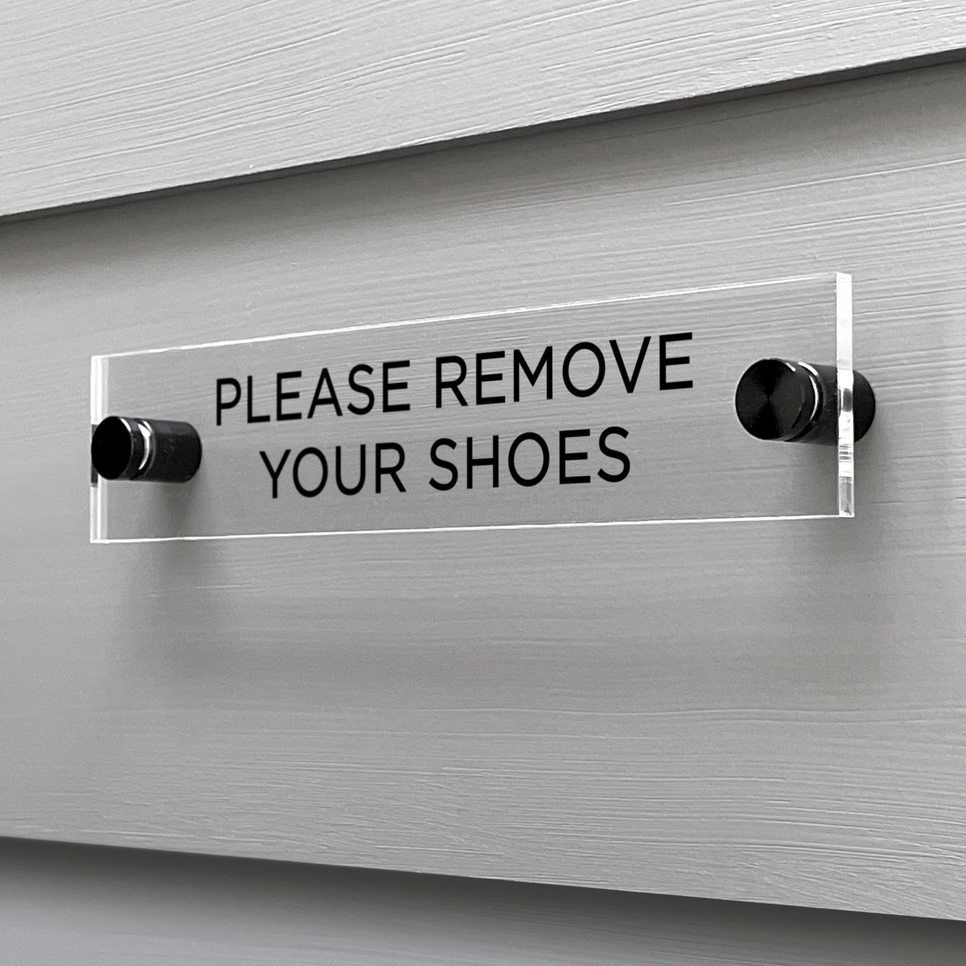 The Girl Friday Shops Please Remove Your Shoes acrylic sign displays bold, black letters and is elegantly mounted on a light gray, horizontally-grooved wall with two metal supports.