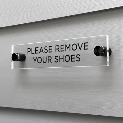 The Girl Friday Shops Please Remove Your Shoes acrylic sign displays bold, black letters and is elegantly mounted on a light gray, horizontally-grooved wall with two metal supports.