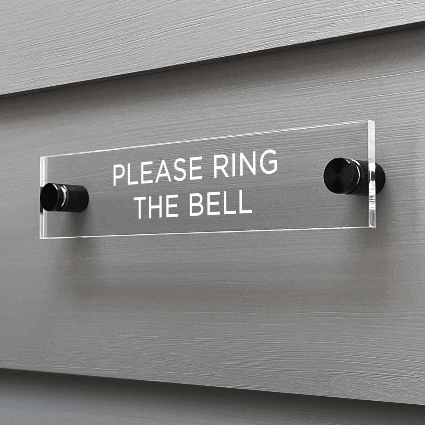 The Girl Friday Shops Please Ring The Bell Acrylic Sign is mounted on a gray wall. This clear, rectangular sign features white capital letters and is securely attached with two black knobs.