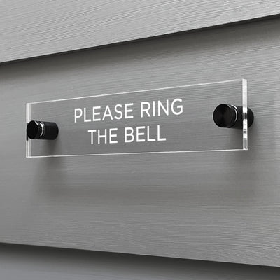 The Girl Friday Shops Please Ring The Bell Acrylic Sign is mounted on a gray wall. This clear, rectangular sign features white capital letters and is securely attached with two black knobs.