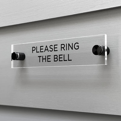 The Girl Friday Shops Please Ring The Bell acrylic sign, featuring black text, is elegantly mounted on a gray wall with metal standoffs.