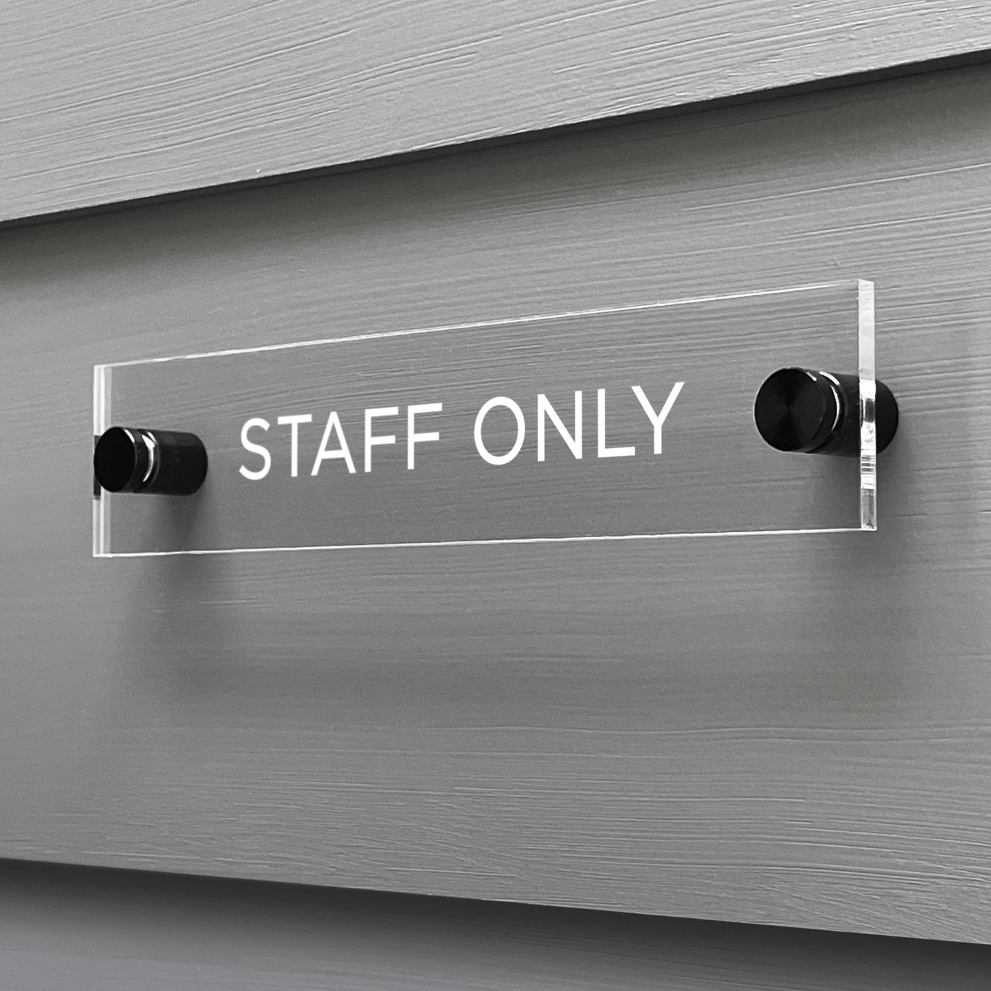 The Girl Friday Shop custom-designed Staff Only Acrylic Sign features crisp white letters on a sleek background, mounted with black attachments and securely affixed to a gray wall.