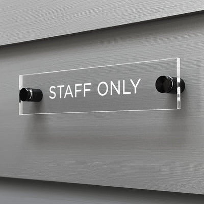 The Girl Friday Shop custom-designed Staff Only Acrylic Sign features crisp white letters on a sleek background, mounted with black attachments and securely affixed to a gray wall.