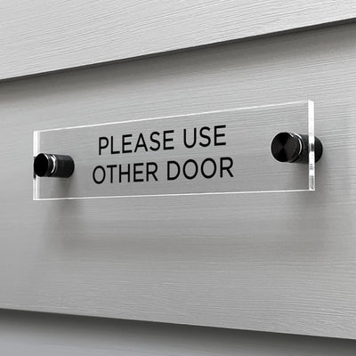 A Please Use Other Door Acrylic Sign by Girl Friday Shop features bold black text on a professionally cut acrylic, mounted on a gray wall.