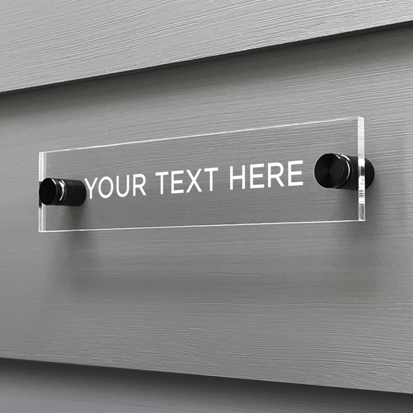 A clear acrylic sign by Girl Friday Shop is mounted on a grey wall with two black screws. Featuring YOUR TEXT HERE in white capitals, the Design Your Own Acrylic Sign invites you to personalize with custom text for your message area.