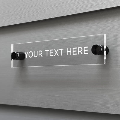 A clear acrylic sign by Girl Friday Shop is mounted on a grey wall with two black screws. Featuring YOUR TEXT HERE in white capitals, the Design Your Own Acrylic Sign invites you to personalize with custom text for your message area.