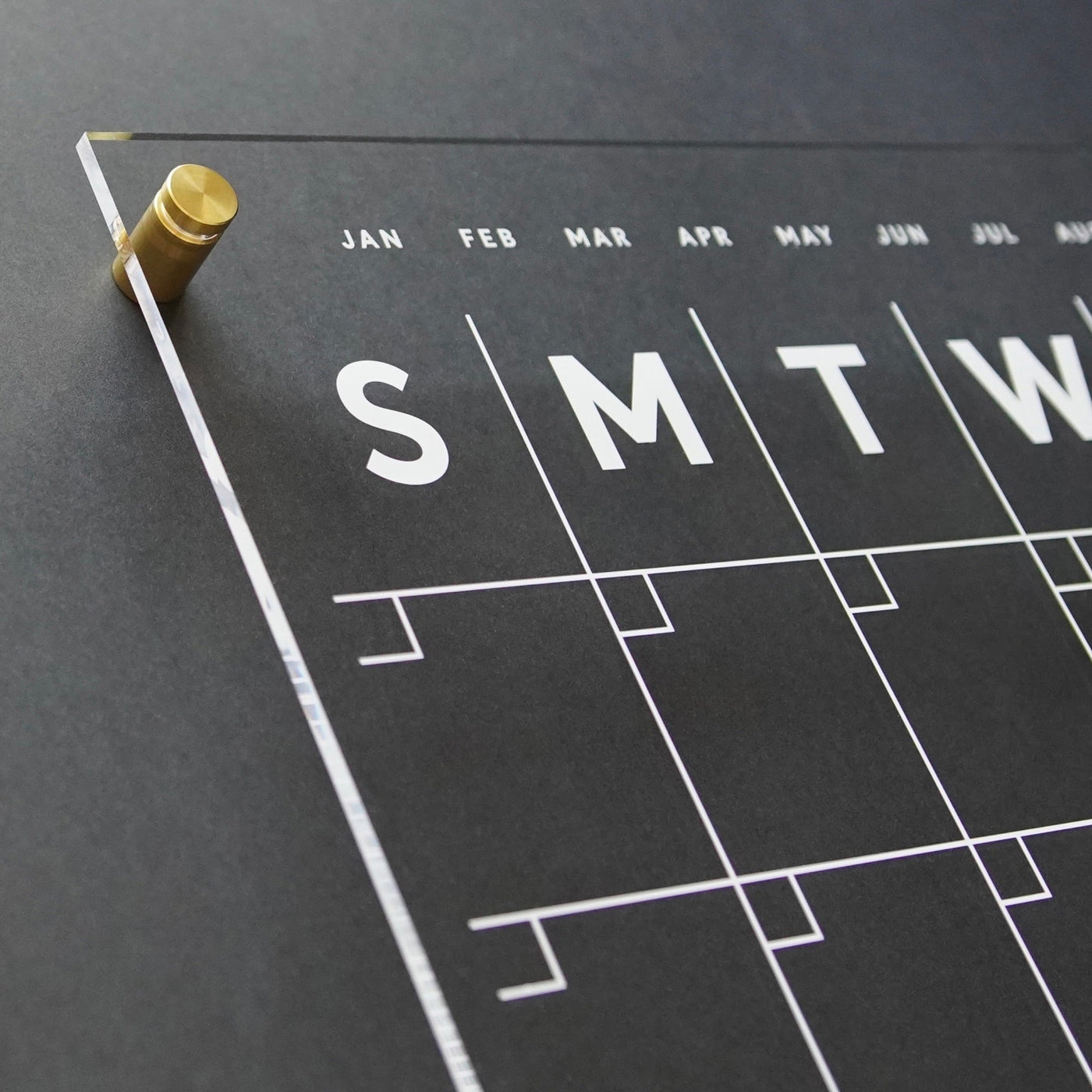 A close-up showcases the Acrylic Calendar with side notes, featuring a modern black and white design with brass pegs. Large initials for days, minimalist date lines, and month abbreviations grace its sleek writing surface—ideal for pairing with wet-erase markers. Brand: Girl Friday Shop.