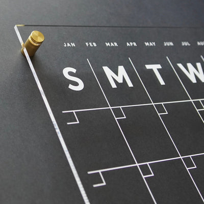 A close-up showcases the Acrylic Calendar with side notes, featuring a modern black and white design with brass pegs. Large initials for days, minimalist date lines, and month abbreviations grace its sleek writing surface—ideal for pairing with wet-erase markers. Brand: Girl Friday Shop.