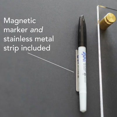 A magnetic marker with a black cap is held by a stainless metal strip against dark walls. White text on the left reads, Magnetic marker and stainless metal strip included, crafted from high-quality materials. Product: Clear Acrylic Monthly Wall Calendar | Horizontal | Girl Friday Shop.
