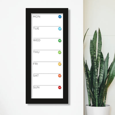 A 9 X 24 weekly whiteboard calendar by CalendarGirlCompany, with a premium acrylic surface, hangs on a white wall. It displays Monday to Sunday, adorned with colorful dots. Nearby, a potted snake plant brings a touch of nature.