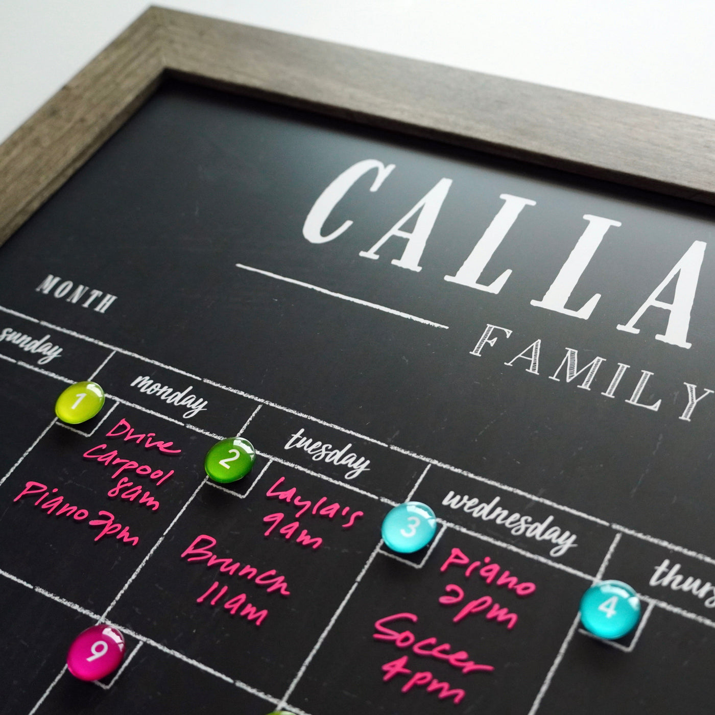 A close-up of the 2025 Personalized Dry Erase Framed Monthly 18 x 24 Calendar by CalendarGirlCompany shows days marked with colorful round magnets and activities like carpool, piano, and brunch written in bright pink ink. The calendar has a black background, but the top text is partially cut off.
