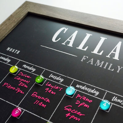 A close-up of the 2025 Personalized Dry Erase Framed Monthly 18 x 24 Calendar by CalendarGirlCompany shows days marked with colorful round magnets and activities like carpool, piano, and brunch written in bright pink ink. The calendar has a black background, but the top text is partially cut off.