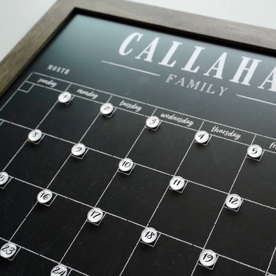 The CalendarGirlCompany 24 X 18 Framed Chalkboard Calendar (#18210) has a customizable frame with a monthly grid. Days are marked by numbered white circles, and Callahan Family is written at the top. Its ideal for use with colorful chalk ink markers.