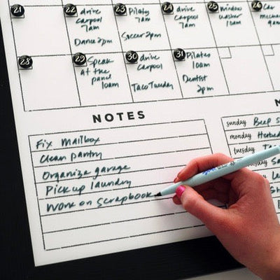 A pen writes Work on Scrapbook on the #18210 Framed Whiteboard Calendar by CalendarGirlCompany. Tasks like fixing the mailbox, cleaning the pantry, and organizing the garage are listed. The premium acrylic surface is filled with colorful reminders using chalk ink markers.