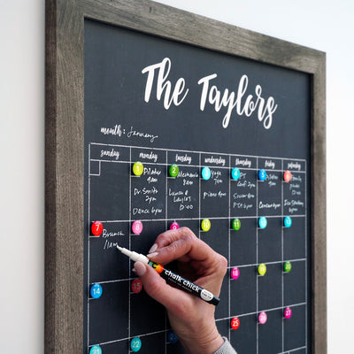 A hand writes on the CalendarGirlCompany 24 X 18 Framed Chalkboard Calendar adorned with the title The Taylors. Colorful magnets are placed around a customizable frame showcasing a weekly January layout filled with appointments and events in vibrant chalk ink markers.