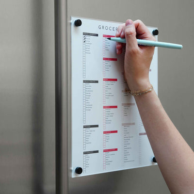A person uses a teal pen and wears a gold bracelet while checking off items on a list under the Girl Friday Shops Magnetic Chore Chart cover, which is attached to a metallic surface. The list is organized by categories with checkboxes.