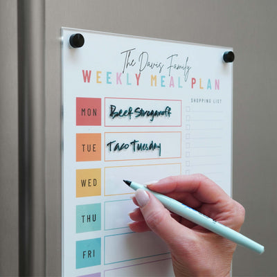 Using the Magnetic Chore Chart cover by Girl Friday Shop, a person writes on an attached magnetic dry-erase weekly meal plan on the fridge. Bright sections mark each day, with Beef Stroganoff and Taco Tuesday noted for Monday and Tuesday. A handy shopping list section is conveniently on the right.