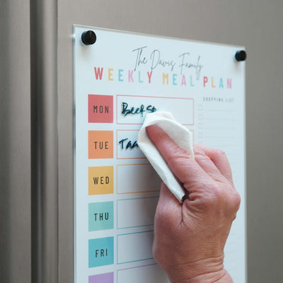 A hand wipes text from Girl Friday Shops Magnetic Chore Chart cover on a metal board showing color-coded days, Monday through Wednesday. A shopping list section is on the right, allowing easy reuse.