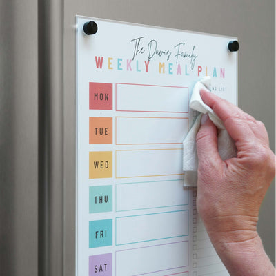 A person wipes a magnetic dry erase board on the fridge, using the Magnetic Chore Chart cover by Girl Friday Shop. Titled Weekly Meal Plan, it shows rainbow-colored days and a side for shopping lists, enhancing meal planning efficiency.