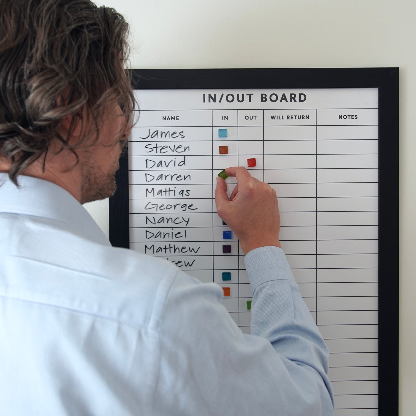 Someone in a light blue shirt updates the Girl Friday Shop 18 X 24 Framed In/Out Magnetic Staff Board, which has a premium acrylic surface. It is customizable with vertical names and In, Out, and Will Return columns, using colorful magnets to indicate each persons status.