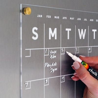 A hand uses a white chalk marker to write Yoga 6am and Playdate 3pm on the Girl Friday Shops Clear Acrylic Monthly Fridge Calendar. The magnetic surface neatly displays the days, with a metallic background gleaming and a gold fastener keeping everything secure.