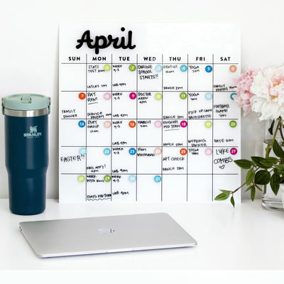 The White Acrylic Monthly Calendar by Girl Friday Shop for April is adorned with colorful notes on a white surface. To its left is a teal travel mug, a closed silver laptop sits in front, and a vase of white flowers on the right provides decoration.