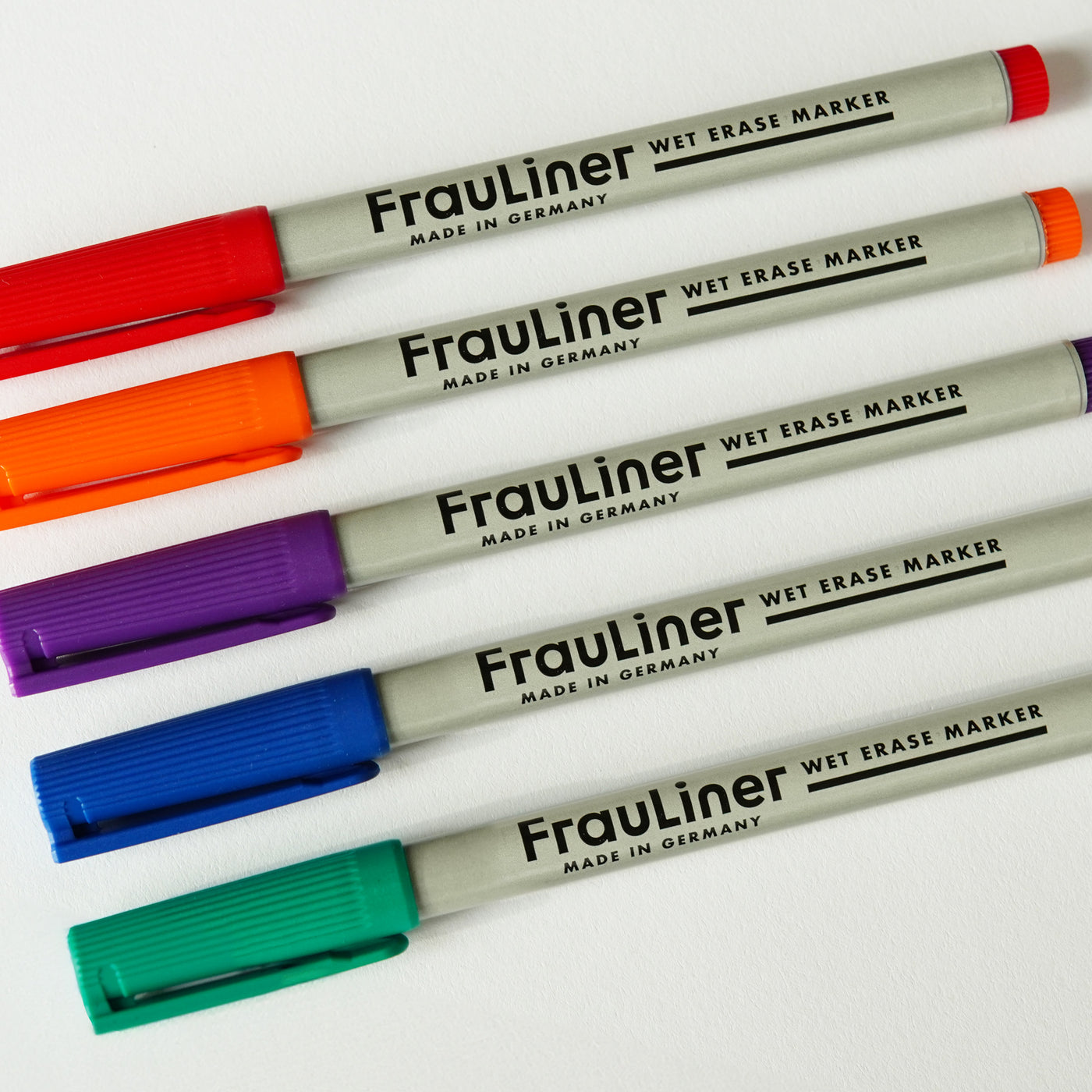 Five FrauLiner wet-erase markers from Girl Friday Shop, with vibrant caps in orange, red, purple, blue, and teal, are labeled Made in Germany and neatly lined up on a white surface. These fine-tip markers are non-toxic and work perfectly on acrylic boards.