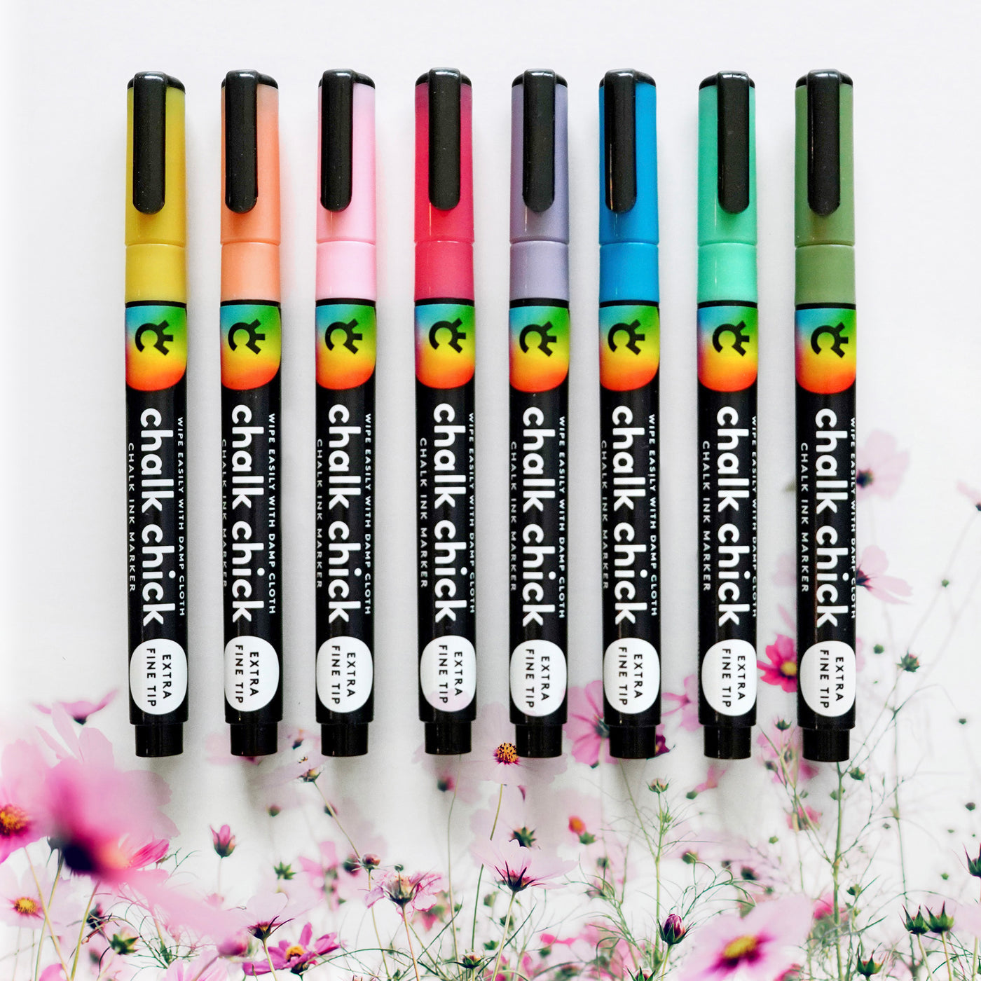 Eight Floral Chalk Ink Markers from Girl Friday Shop, featuring extra fine tips, stand vertically in colorful hues—black to purple, pink, red, and blue—on a white surface. Floral designs are partially visible on the markers, perfect for use on acrylic boards.