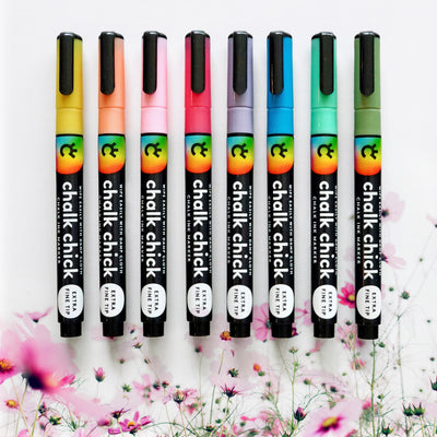 Eight Floral Chalk Ink Markers from Girl Friday Shop, featuring extra fine tips, stand vertically in colorful hues—black to purple, pink, red, and blue—on a white surface. Floral designs are partially visible on the markers, perfect for use on acrylic boards.