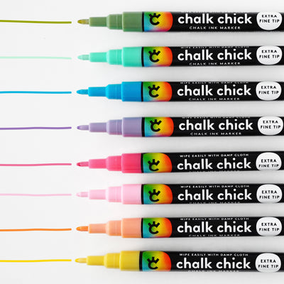 Ten vibrant Floral Chalk Ink Markers by Girl Friday Shop, perfect for acrylic boards, line up horizontally, each leaving a matching line above. With extra fine tips and black bodies labeled Wipe Easily with Damp Cloth, colors range from yellow to pink and purple.