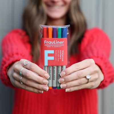 A person in a red sweater holds a pack of colorful markers labeled Made in Germany towards the camera. Their face is out of focus, and they wear several rings on their fingers—ideal for adding flair to your Girl Friday Design Your Own Floating White Acrylic Board | Vertical.