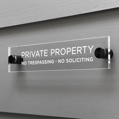 A Private Property Acrylic sign by Girl Friday Shop, featuring the text NO TRESPASSING · NO SOLICITING, is mounted on a gray wall using two black fasteners.