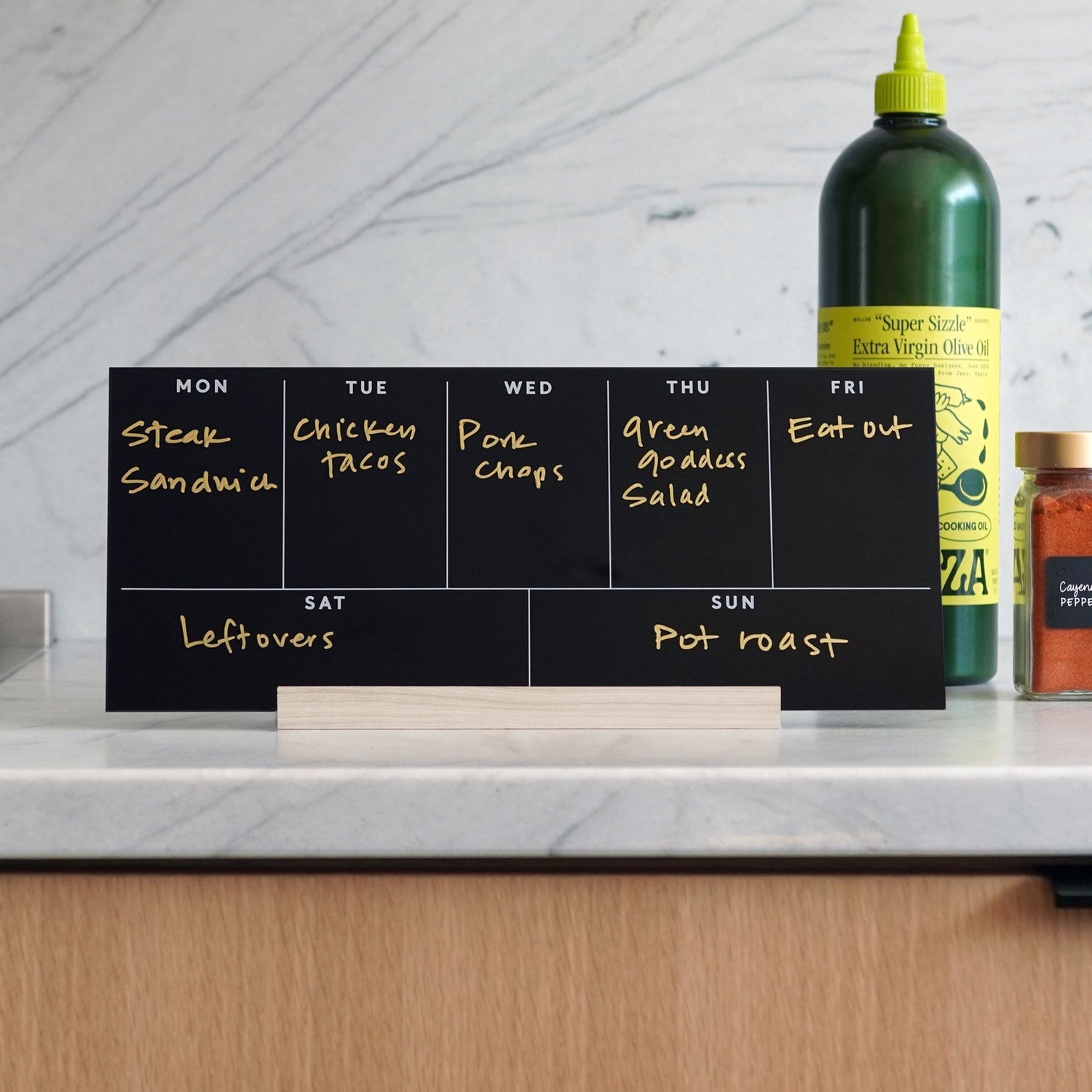 The Girl Friday Weekly Black Acrylic Calendar with Wood Stand doubles as a meal planner, displaying meals like steak sandwich and chicken tacos. A large green bottle and spice jar on the kitchen counter enhance this organized culinary setup.