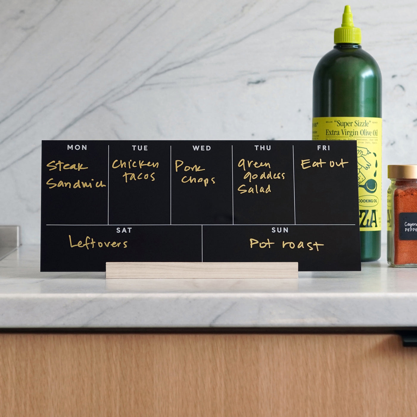 The Girl Friday Weekly Black Acrylic Calendar with Wood Stand doubles as a meal planner, displaying meals like steak sandwich and chicken tacos. A large green bottle and spice jar on the kitchen counter enhance this organized culinary setup.