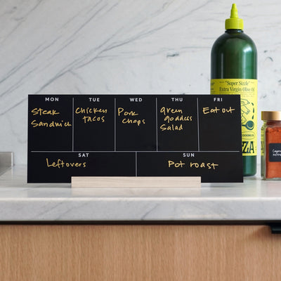 The Girl Friday Weekly Black Acrylic Calendar with Wood Stand doubles as a meal planner, displaying meals like steak sandwich and chicken tacos. A large green bottle and spice jar on the kitchen counter enhance this organized culinary setup.
