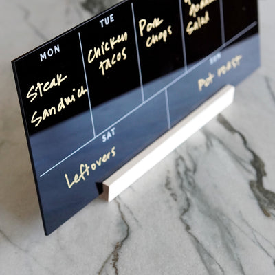 The Weekly Black Acrylic Calendar with Wood Stand by Girl Friday doubles as a sleek tabletop menu planner on a marble surface, displaying meals: Monday - Steak Sandwich, Tuesday - Chicken Tacos, Wednesday - Pork Chops, Thursday - Enchiladas, Friday - Salad, Saturday - Leftovers, Sunday - Pot Roast.