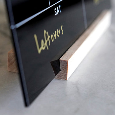 A close-up showcases the Weekly Black Acrylic Calendar with Wood Stand by Girl Friday, partially displaying Leftovers in yellow under SAT. This sleek, reusable acrylic planner is perfect for weekly reminders, resting stylishly on a gray countertop.